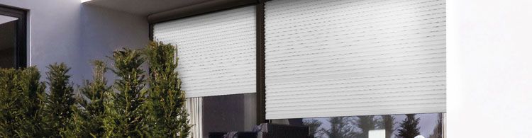 Zwei große Fenster mit heruntergelassenen weißen Rollläden an einer modernen Hausfassade, davor grüne Büsche.